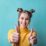 Happy woman in yellow giving thumbs up against a teal background, expressing positivity.