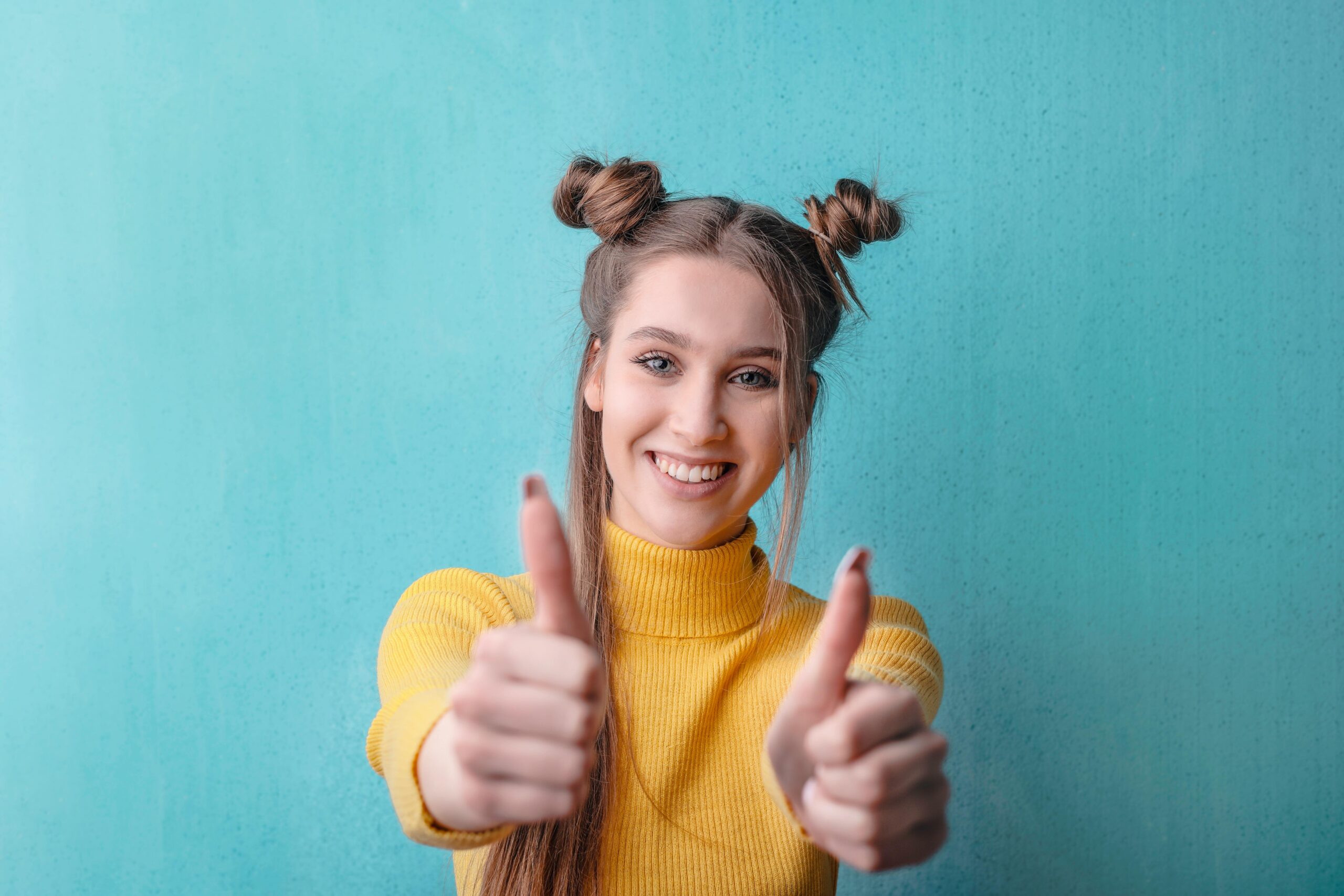 Happy woman in yellow giving thumbs up against a teal background, expressing positivity.