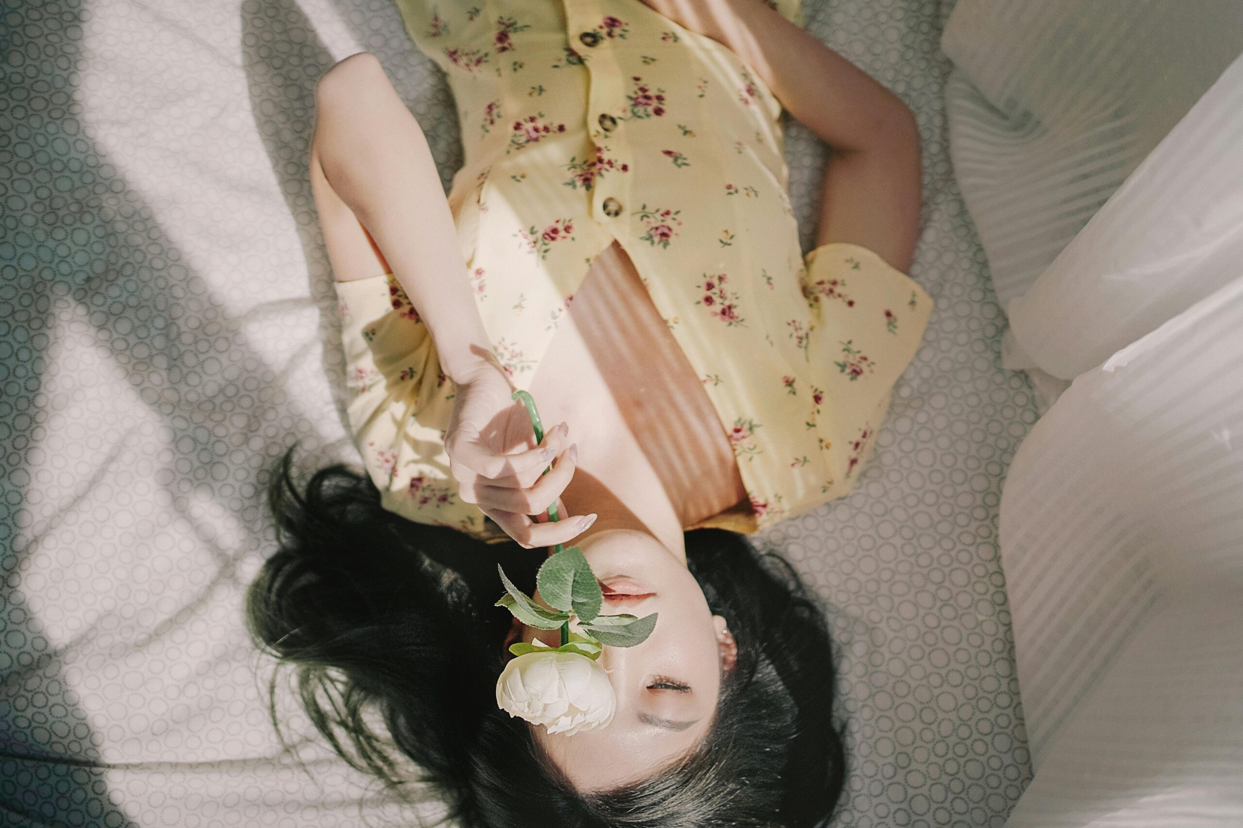 Young woman in floral dress holding a rose, relaxed on bed in morning light.