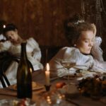 Two women in period costumes asleep at a candle-lit dinner table, evoking opulence and historical charm.