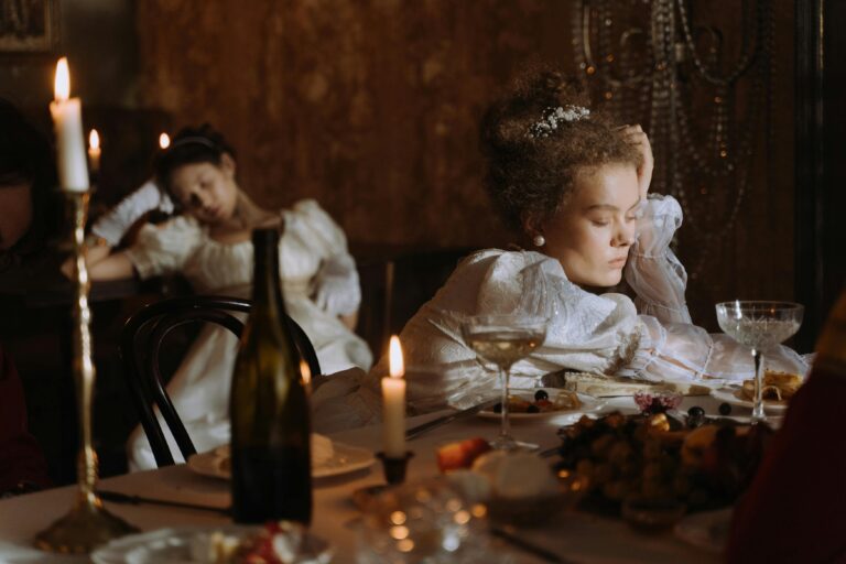 Two women in period costumes asleep at a candle-lit dinner table, evoking opulence and historical charm.