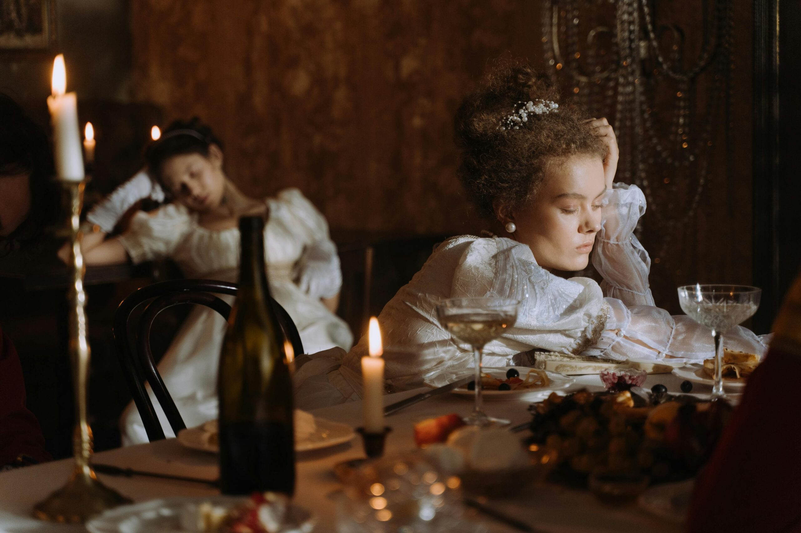 Two women in period costumes asleep at a candle-lit dinner table, evoking opulence and historical charm.